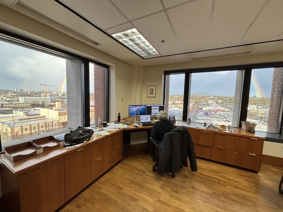 Commissioner Meghan Moyer working at her desk with a view of the ends of rainbow over the city scape view outside her windows. on the sixth floor of the Multnomah Building.