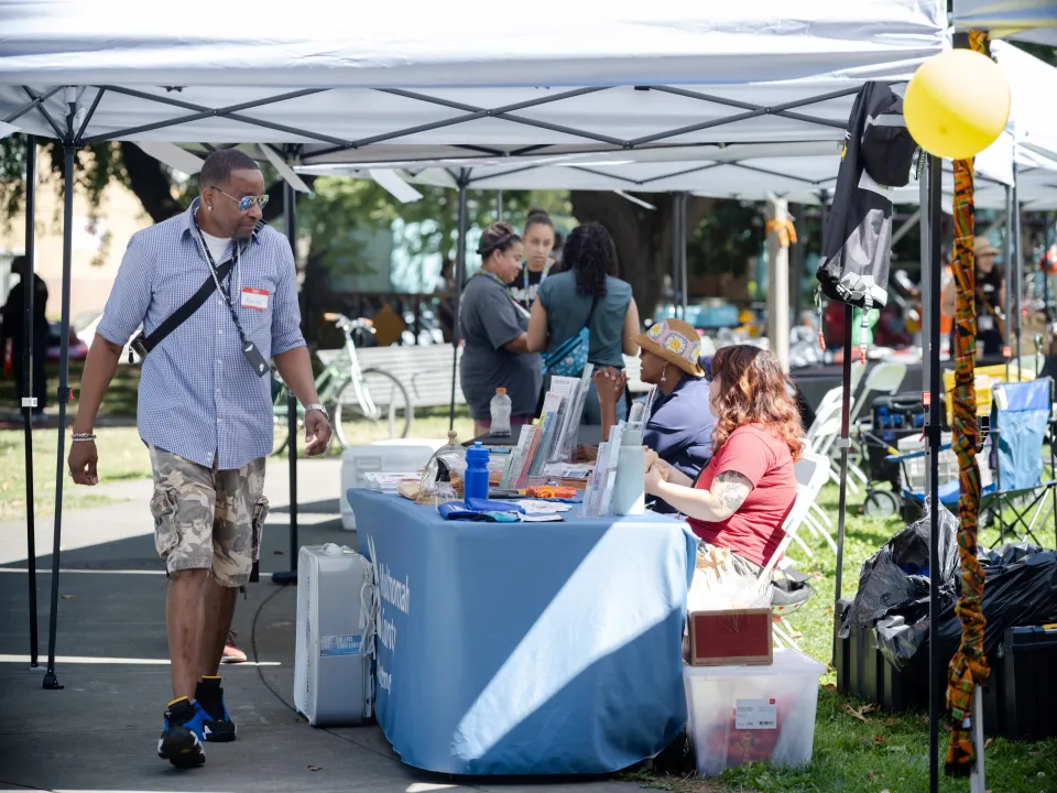 A tall Black man with sunglasses walking by a table under a tent at a fair