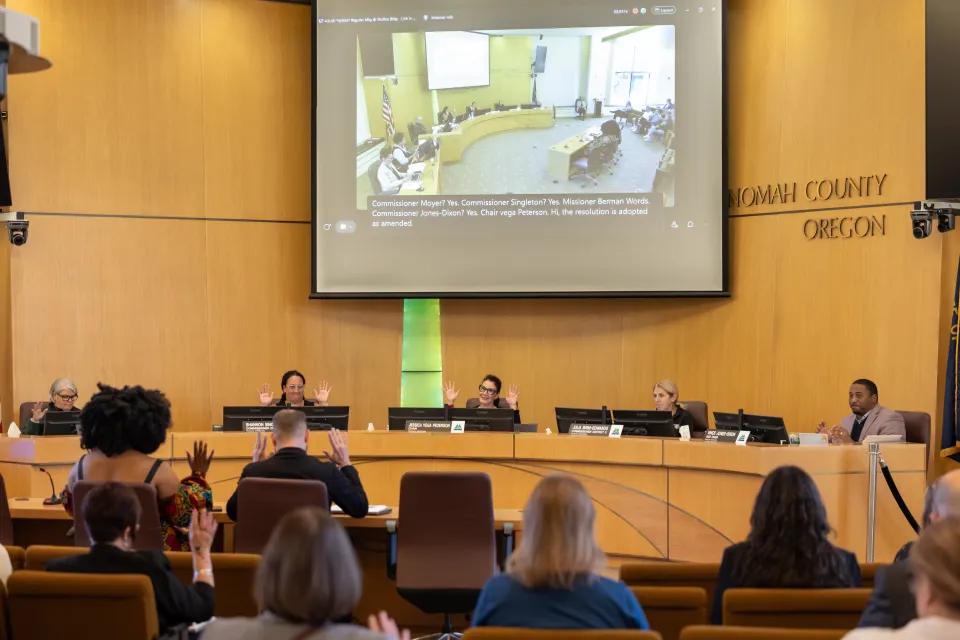 The Multnomah County Board of Commissioners and members of the public raise their hands in a unanimous vote during a board meeting