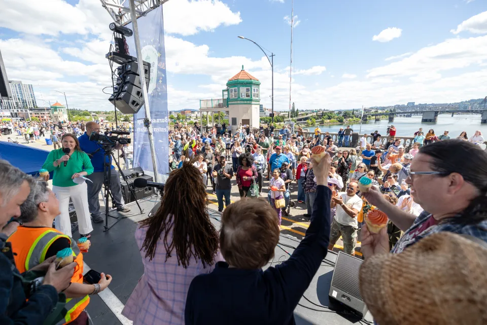 A crowd gathers around a stage on the Burnside Bridge with the bridge towers visible in the background. The bridge is closed to cars and packed with celebrating people.