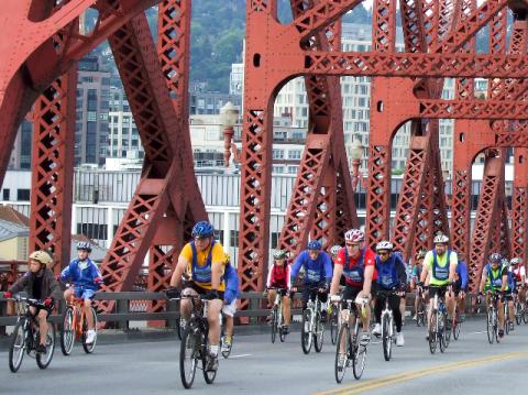 Large number of people on bikes filling the two eastbound lanes of the Broadway Bridge, with distinctive red girders around them and the Pearl District in the background.