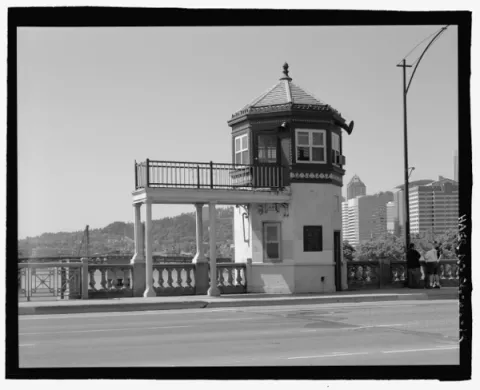Black and white image of the bridge operator's house on the Burnside Bridge, with parts of downtown and Marquam Hill in background. House is two stories with a cupola on top and a long portico supported pillars emerging from the second floor.