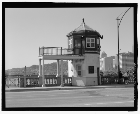 Black and white image of the bridge operator's house on the Burnside Bridge, with parts of downtown and Marquam Hill in background. House is two stories with a cupola on top and a long portico supported pillars emerging from the second floor.