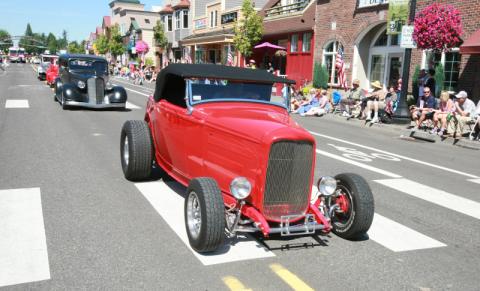 A red roadster leads a parade of classic cars through downtown Troutdale, with a crowd sitting on the sidewalk with an American flag.