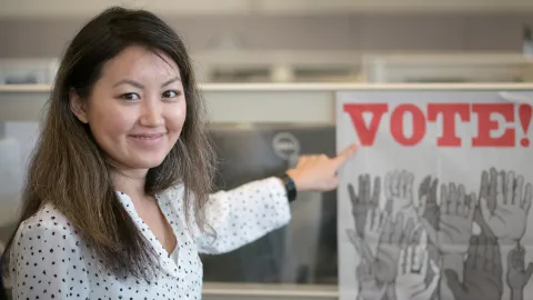Elections worker smiles and points to a VOTE poster at the elections office. Poster shows many hands raised.