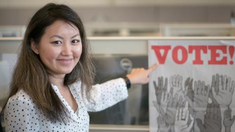 Elections worker smiles and points to a VOTE poster at the elections office. Poster shows many hands raised.