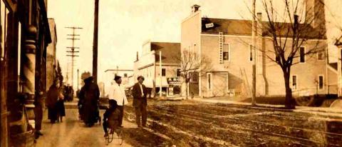 A grainy, sepia-toned image of people in old-fashioned clothes standing next to a muddy street.