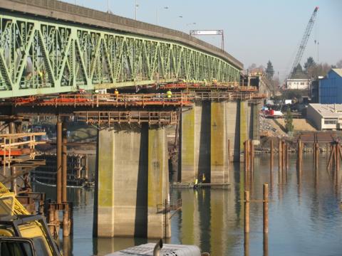 The main deck of the old Sellwood Bridge, raised on hydraulic jacks above its support towers, with workers in bright safety vests looking tiny in comparison.