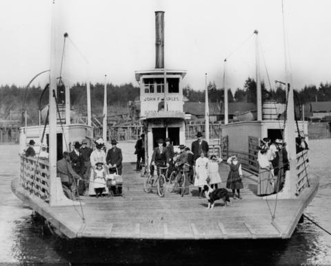 Black and white photo of a steam ferry carrying people in old-fashioned clothing, two bicyclists and a dog.