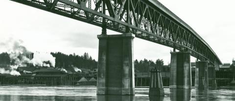 A black and white photo looking up at the sellwood bridge from river level, with factories and smokestacks on the far riverbank.