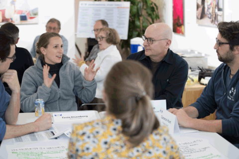 A woman seated at a table, gesturing with her hands as she speaks, while others around the table listen intently.