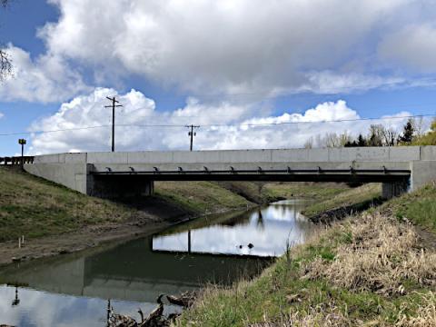 A concrete bridge over a narrow creek, with its own reflection in the water below and clouds in a blue sky above.