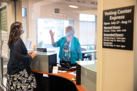 Elections staff wearing a face mask and behind plexiglass waves to voter wearing a face mask that she is helping at the Voting Center Express in Gresham