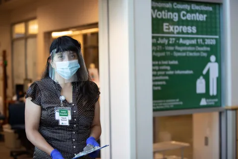 Elections staff wearing a face shield, face mask, and gloves stands in front of the Voting Center Express in Gresham. She is holding a clipboard and wears an "I Speak Spanish / Hablo Español" badge