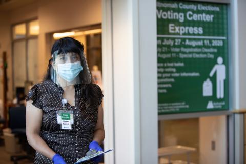 Elections staff wearing a face shield, face mask, and gloves stands in front of the Voting Center Express in Gresham.  She is holding a clipboard and wears an "I Speak Spanish / Hablo Español" badge