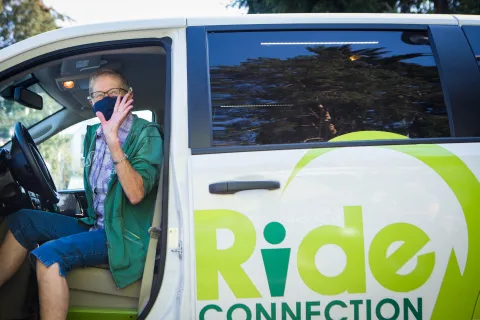 A driver wearing a cloth mask waves from the front seat of a white van bearing the Ride Connection logo.
