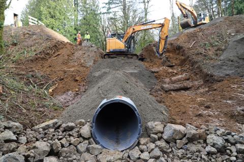 An exposed culvert at the bottom of a slope under construction, with two large excavators at the top.