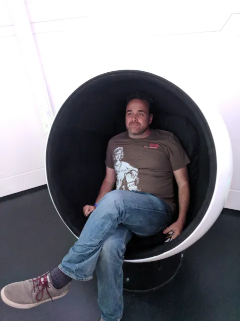 Man in white chair shaped like an egg with one piece cut out. He has stubble on his face, wearing a brown t-shirt blue jeans and canvas shoes.