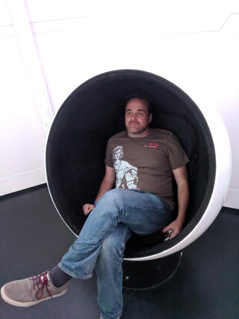 Man in white chair shaped like an egg with one piece cut out. He has stubble on his face, wearing a brown t-shirt blue jeans and canvas shoes. 