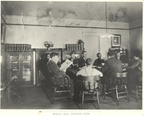 Men and boys sitting around a table reading books and newspapers. Walls of room lined with book shelves/