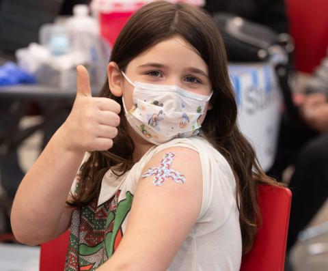 Girl with rainbow bandages on her arm gives a thumbs up to being vaccinated