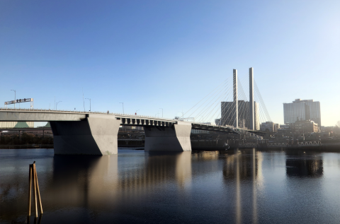 Rendering of a bridge over a river, with two slender columns supporting cables, and tall buildings in the background.