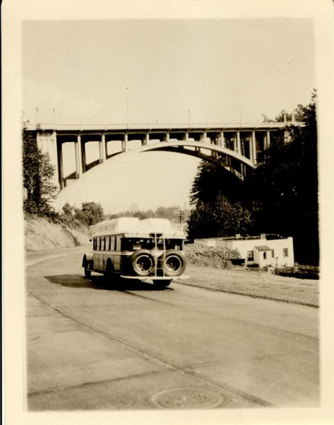 Columbia Gorge System bus passing under Vista bridge on canyon road with a low building off the the right side of the road. The side of the bus reads Columbia Gorge System along the luggage case on top.