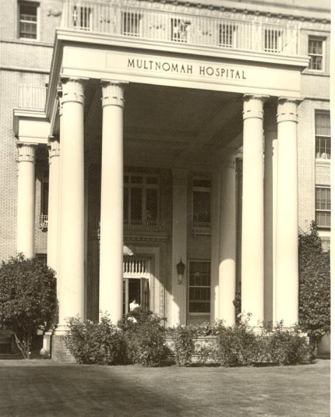 Front portico entrance of multi-story hospital building with someone standing in the open doorway/ Front of portico reads Multnomah Hospital.