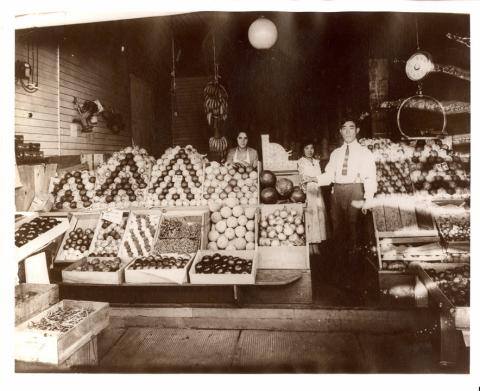 Man and two women standing in the midst of crates of fruit and vegetables placed at the open front of a shop. Scales are visible for weighing.