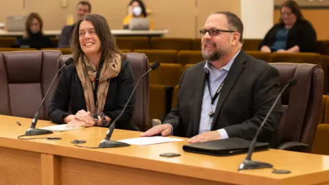 Two people in formal attire sitting at a boardroom table and smiling.