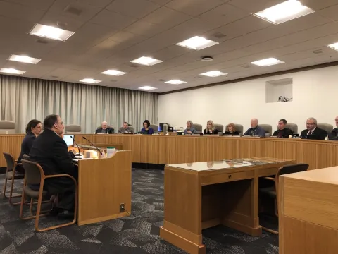 Two people sit at a table facing a panel of questioners in a committee room.