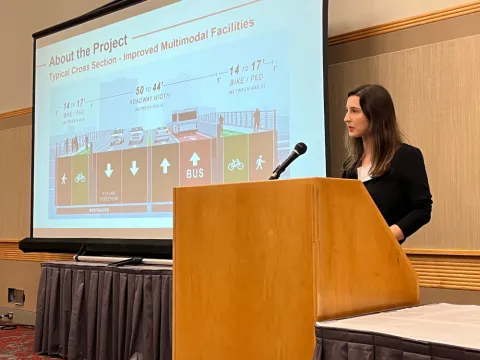 A woman stands at a podium, speaking into a microphone. A screen behind her displays a slide labeled "About the Project - Typical Cross Section - Improved Multi-Modal Facilities".
