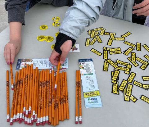 Two children's hands, one with red nail polish and the other with a black wrist brace, reach out to pick up pencils with the Walk and Roll logo, from a pile of pencils on a table surrounded by Walk and Roll stickers of different shapes and sizes.