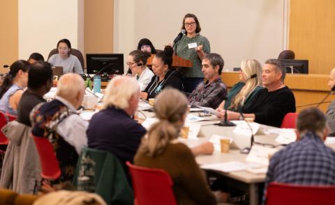 A group of about a dozen people sitting around a long conference table in a board room, with a speaker at a podium beind them.