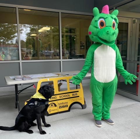 A person in a green and white dragon mascot suit stands next to a black dog with a yellow bandana in front of a sign-up table at a school building.