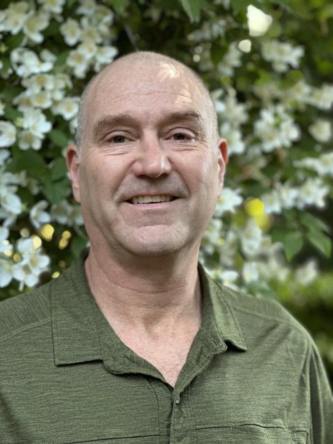 Gideon stands smiling in a green shirt, in front of a wall of green leaves and white-petal flowers.