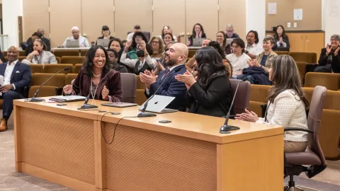 In the foreground, four people sit behind a large table celebrating with applause. In the background is a seated audience looking happy.