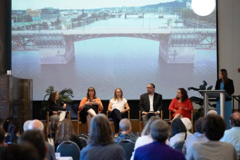 A group of five panelists seated on a stage with a moderator at a podium, facing an audience, with a large image of the Burnside Bridge projected on a screen behind them.