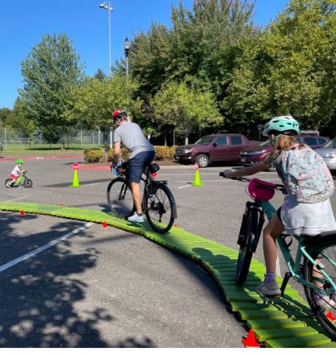 A pair of bicyclists navigate a narrow yellow track in a parking lot, with miniature orange safety cones on the sides.