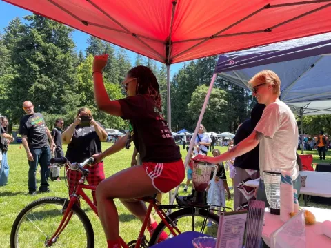 A woman raises a hand as she rides a stationary bike with a blender mounted on the back.