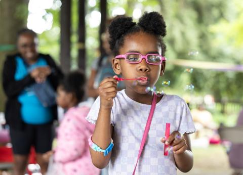 black girl in pink glasses blowing bubbles at a community picnic
