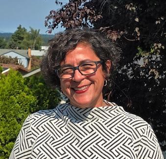 Amara, a light brown skinned middle aged woman with short brown curly hair smiles outside. She is wearing a white shirt with geometric designs and black rimmed glasses. 