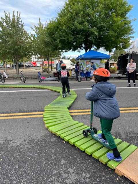 Two kids in bright orange helmets ride scooters along a narrow, bright green scooter track.