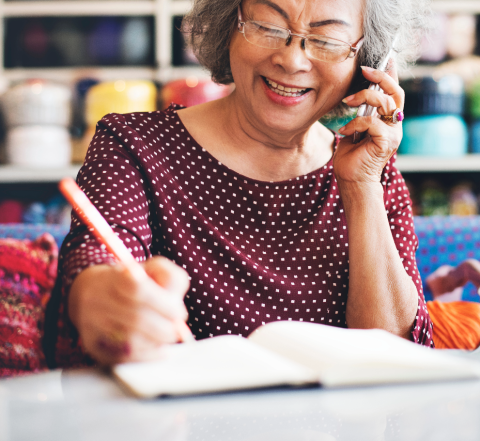 older adult holds a phone up to their ear and pencil, writing notes