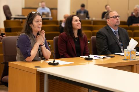Three people in formal attire sit at a presentation table in the board room. They are looking forward and slightly up at some sort of presentation and smiling.