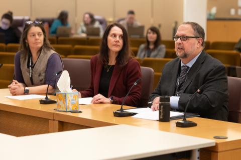 Two people sitting at a presentation table in the board room are looking attentively at a third, who is addressing the commissioners in front of him.