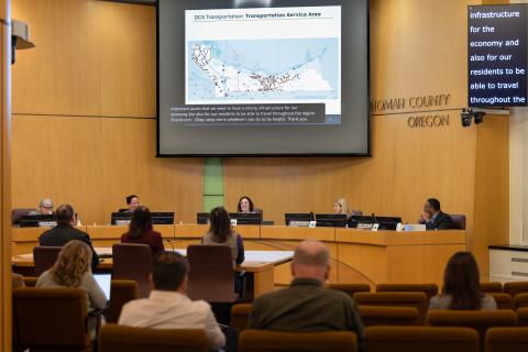 The five board members sit in their seats at the front of the chamber, with the three presenters facing them, and large display above their heads showing a map labeled "DCS Transportation Service Area".