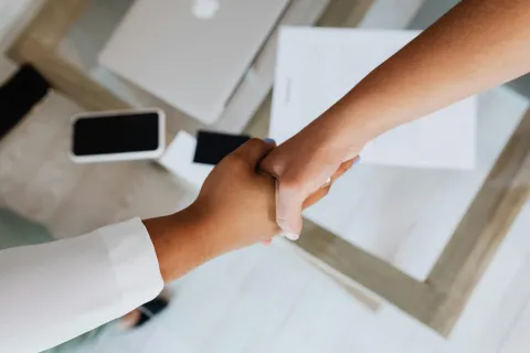Two people handshake over table covered in paper, a laptop, and mobile devices
