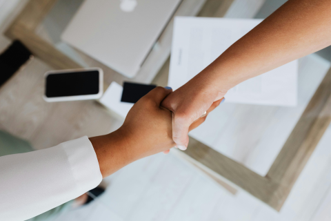 Two people handshake over table covered in paper, a laptop, and mobile devices