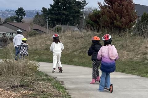 four children riding scooters in single file behind an adult. everyone is wearing a helmet.